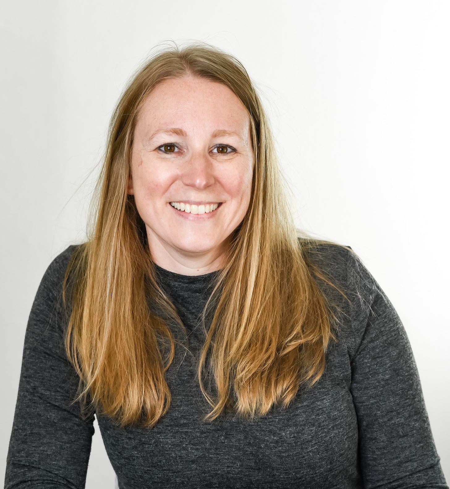 A picture of Meghan Crozier, a white cis blonde woman with long straight hair hanging down past her shoulders. She's wearing a long sleeved, slate gray shirt and smiling.