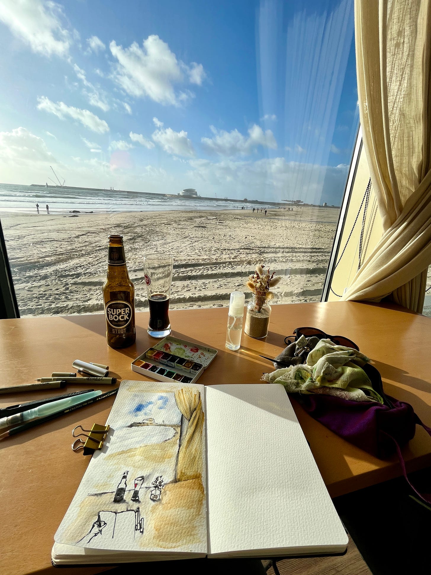 image: loose ink and watercolour sketch on location set up, with a drink, a couple of pens, watercolour kit, overlooking ocean view of blue sky, fluffy white clouds, miles of sand in Matosinhos Beach, Porto. The Port of Leixous in the far horizon can be seen