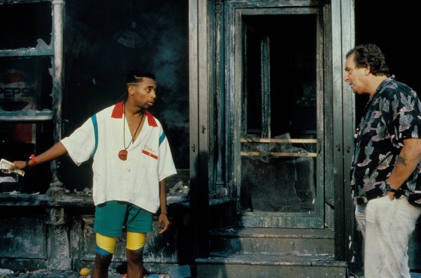 This image shows Mookie (played by Spike Lee, left) and Sal Frangione (played by Danny Aiello, right) standing in front of the shattered windows and burned storefront of Sal's Famous Pizzeria in the final scene of Lee's 1989 film Do the Right Thing. Mookie gestures offscreen, to his right, while Sal stands with his hands in his pants pockets.