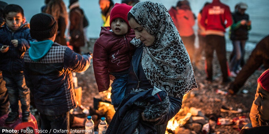 Woman holding child in her arms next to a bonfire on a freezing beach in Greece surrounded by other people