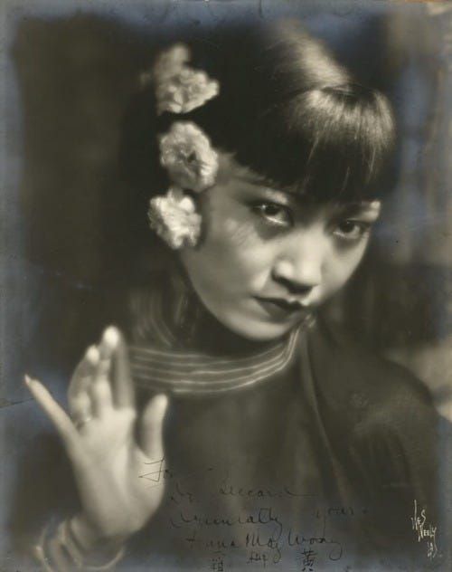 black and white portrait of AMW, she is dressed in Chinese clothing with flowers in her hair, she raises one hand with long fingernails