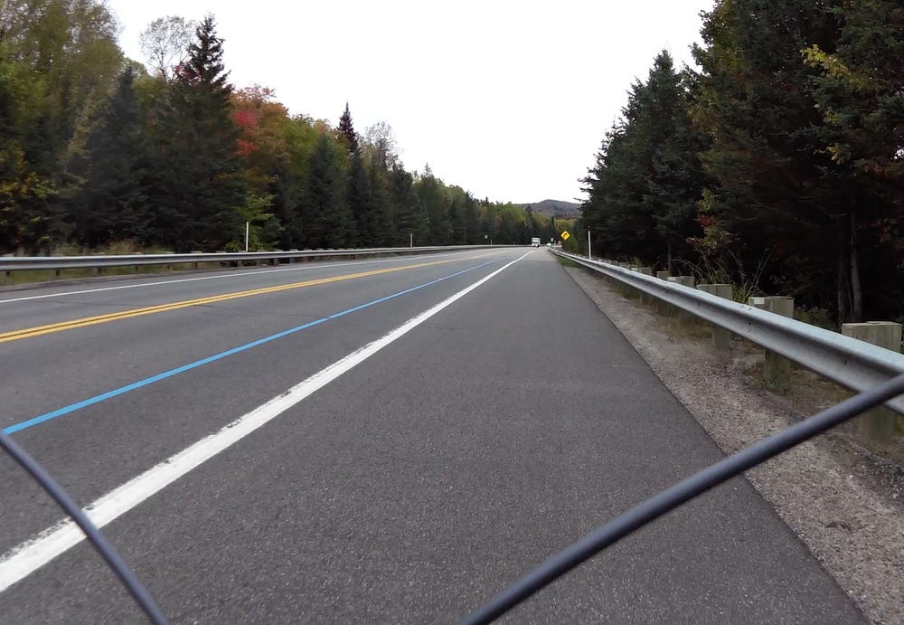 A tree lined highway with a wide shoulder for cycling. A blue line indicates the highway is part of the Iron Man race route.