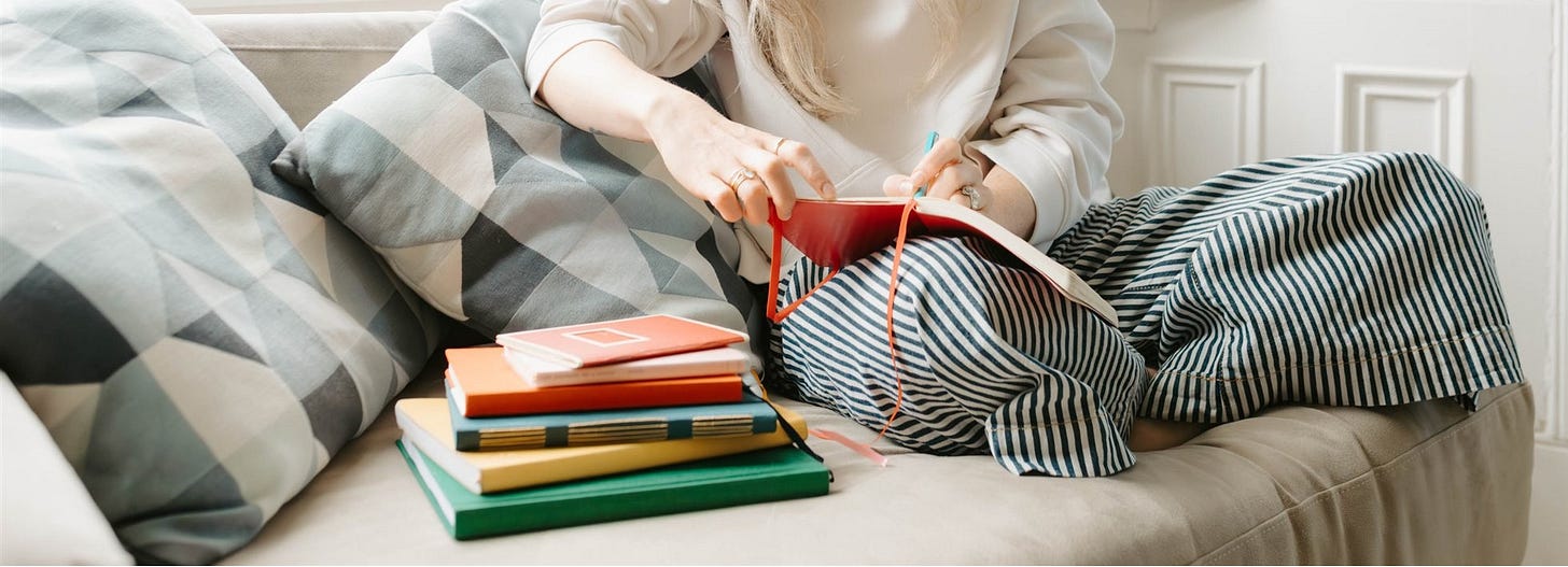 woman sitting on sofa writing, with pile of colourful notebooks beside her