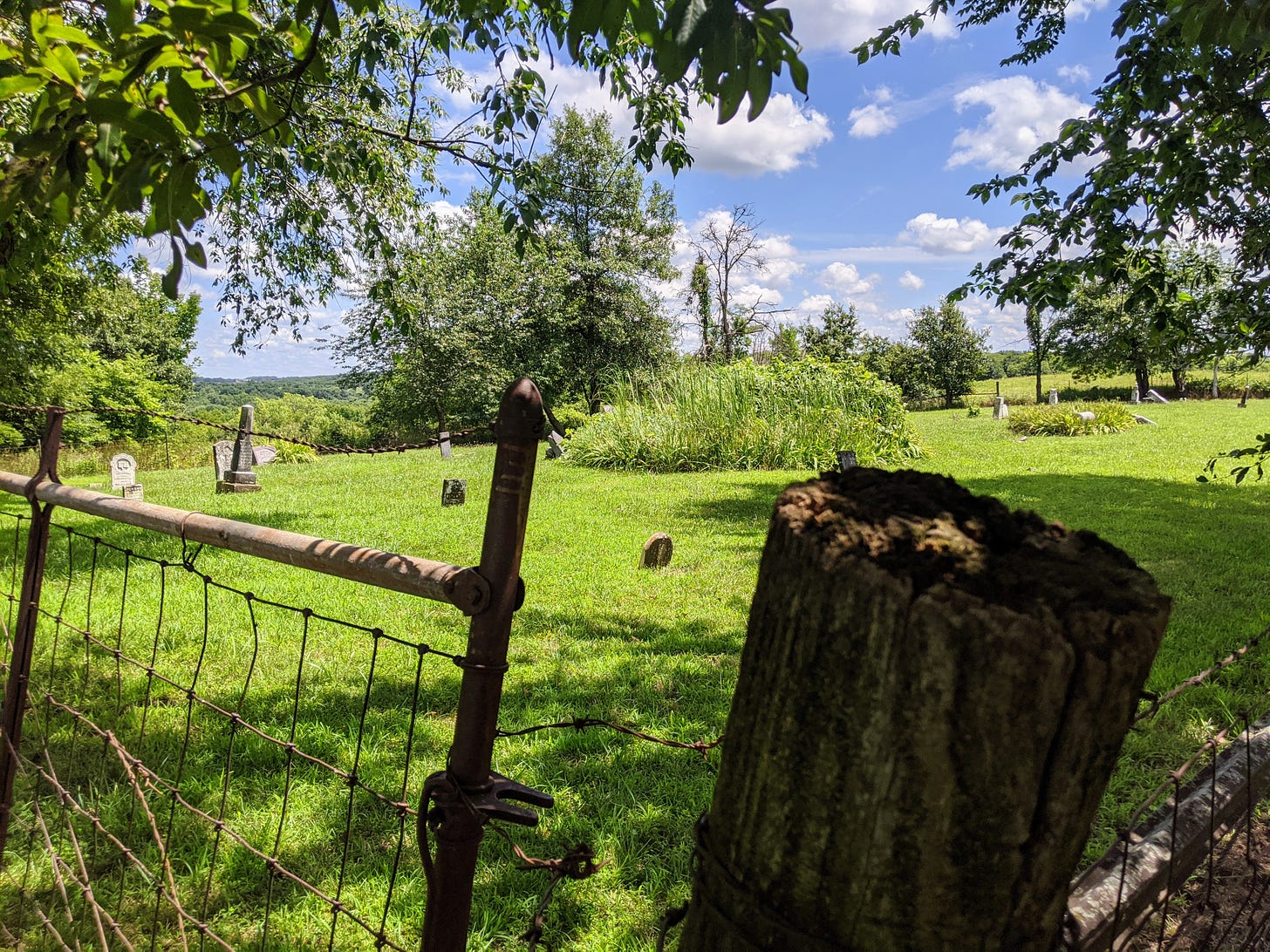 Old cemetery in summer, with a few leaning gravestones, a barbed wire fence, and trees and fields in the distance.