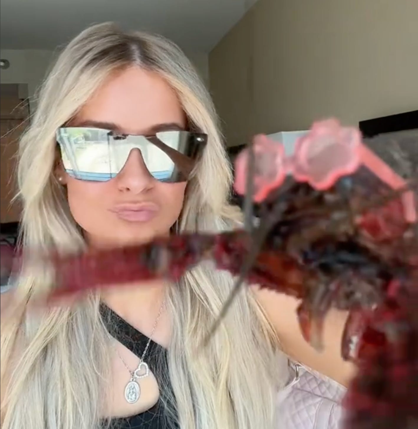 A blonde woman in sunglasses is making duck face while holding up a crawfish to the camera. The crawfish is also wearing sunglasses -- tiny pink flowers.