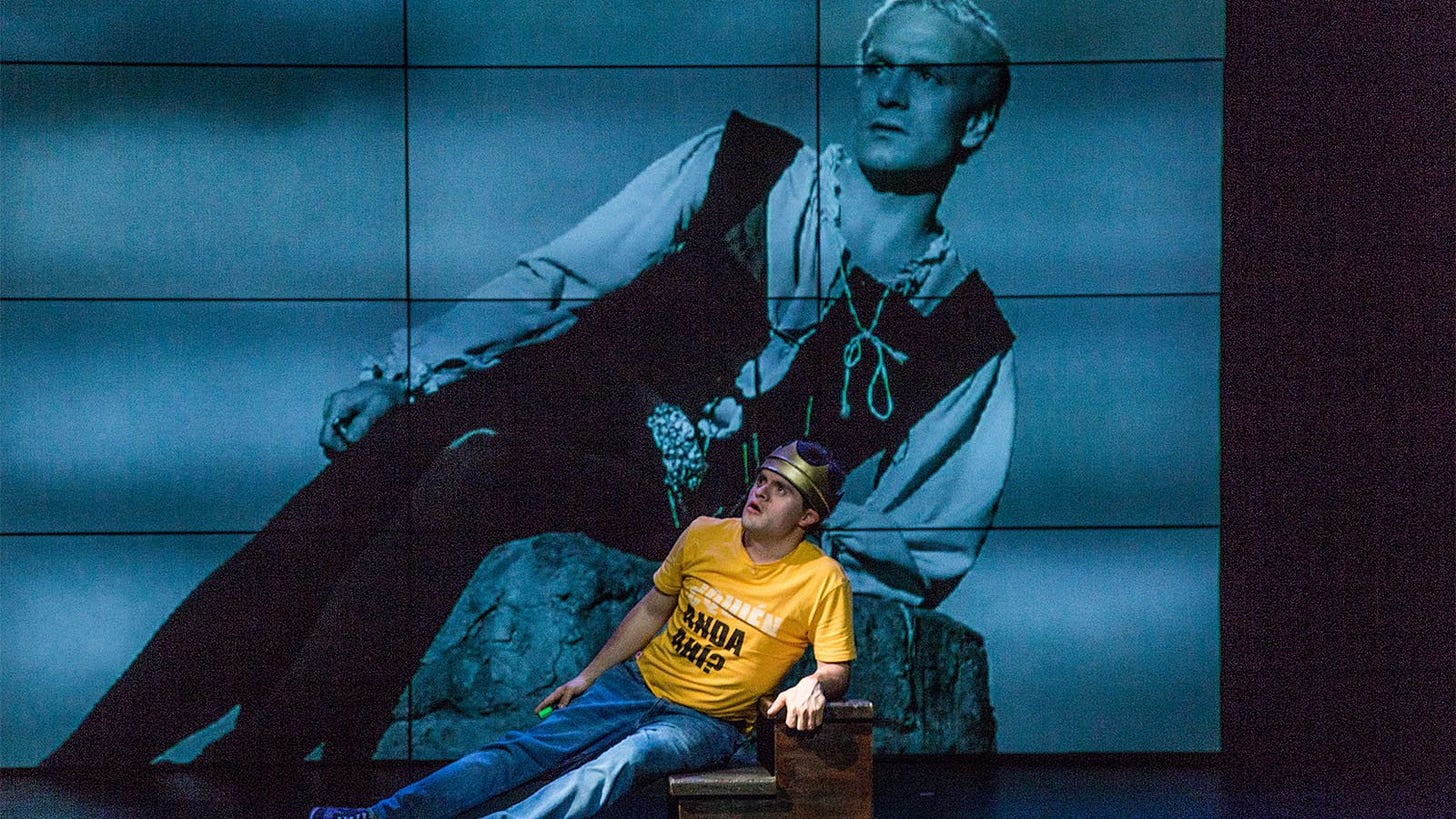 A film still of a black and white film version of Hamlet is on a large screen at the back of a stage. The character is leaning on a rock. The posture is matched by a disabled actor on the stage in front.