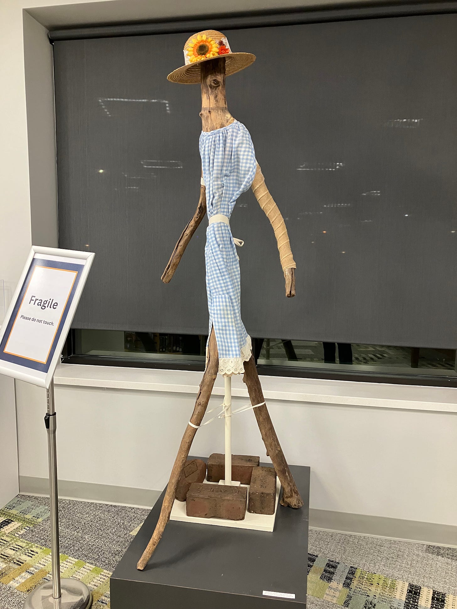 Oak branch sculpture of a tall woman wearing a blue and white gingham dress and a straw hat with artificial flowers