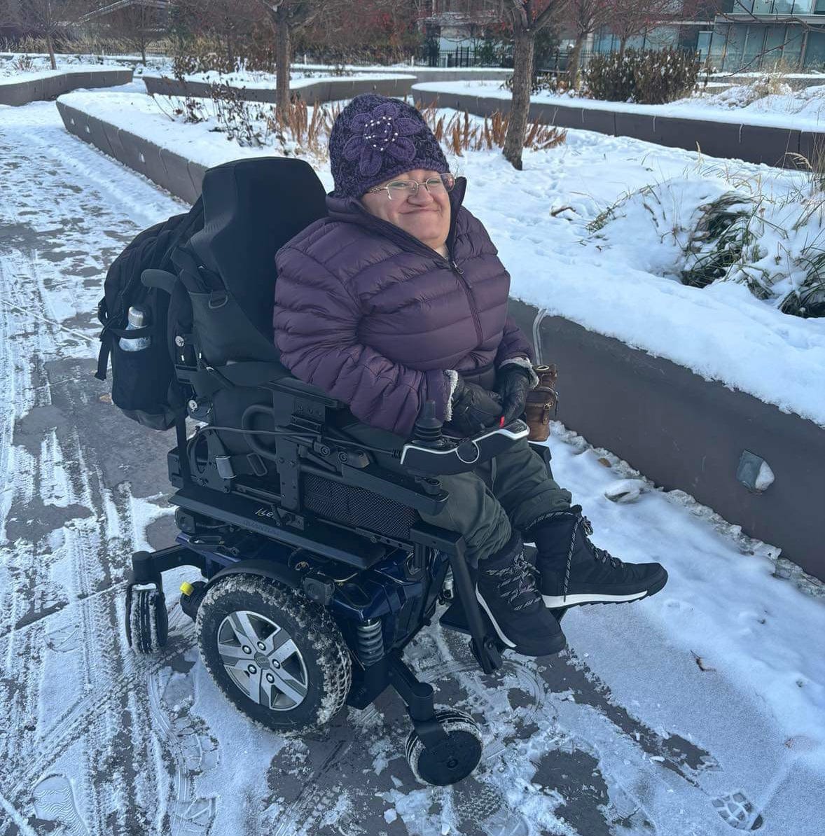 Photo of Athena from last October sitting outside in the patchy snow in her power wheelchair. She's wearing a puffy purple jacket and a matching purple kit toque with a big flower on it.