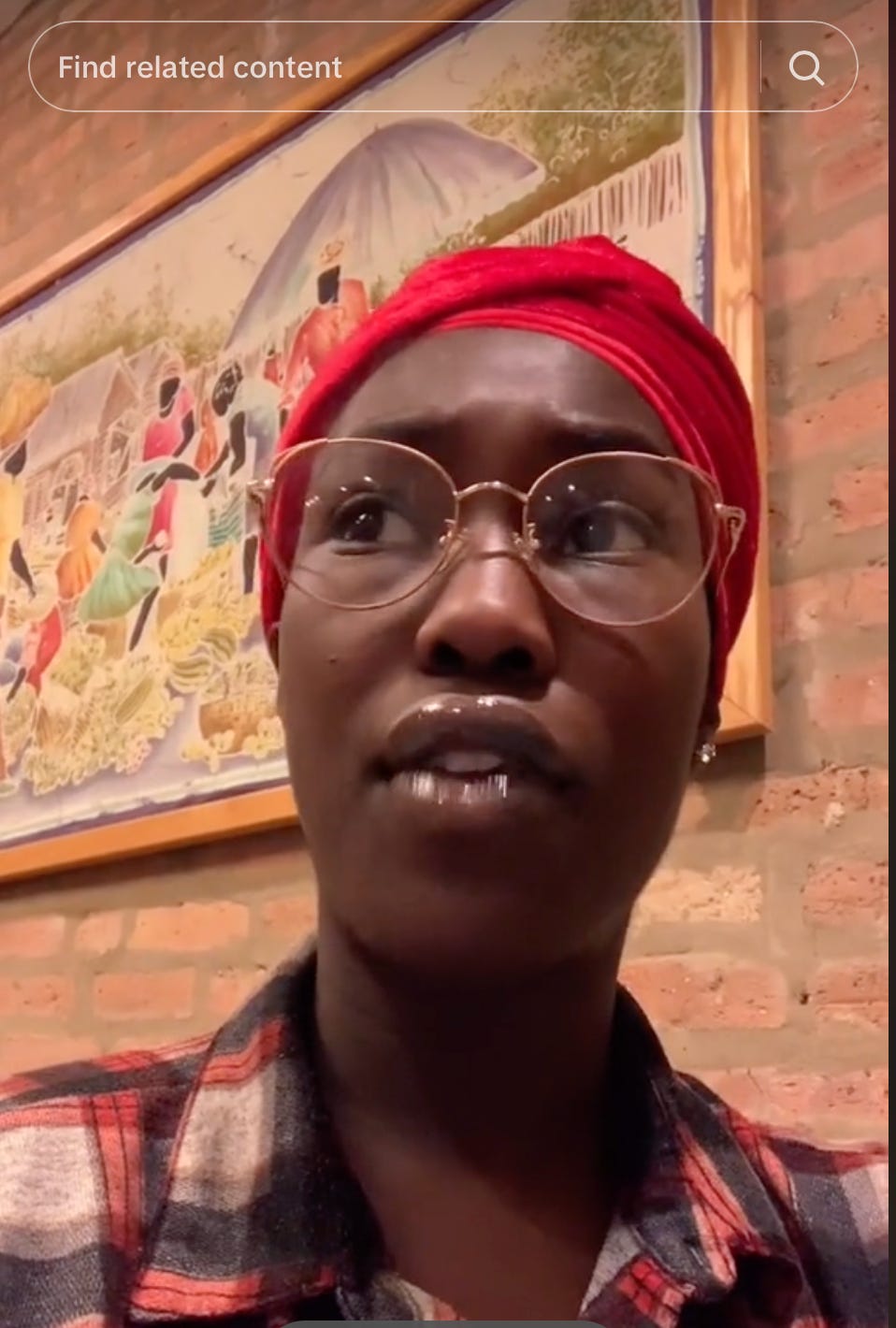 a screenshot of ismatu gwendolyn’s first tiktok; they wear gold wire-rimmed glasses, a glossy lip, and a red velvet headscarf. they are seated in front of a tapestry of an african village on a brick wall.