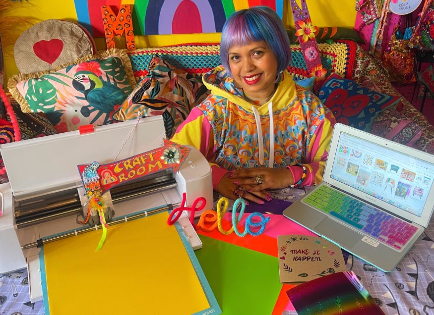 Momtaz with colourful hair and clothing, surrounded by colourful decor and craft supplies, smiling at the camera