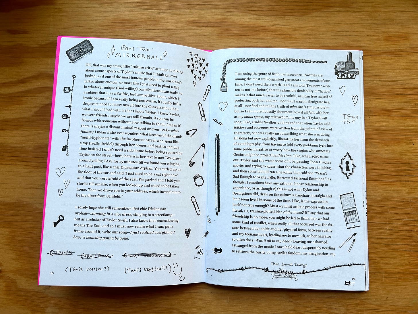 A photograph showing an open copy of the zine “Fan Fiction” laid flat on a wood surface. Two pages of the zine are visible. At the top of the left page, a hand-written header reads “Part Two: Mirrorball.” The text is written in a serif font and the borders of both pages are decorated with miscellaneous images which include doodles of hearts, safety pins, paperclips, matches, charm bracelets. At the bottom of the left page, hand-written doodles read “(Tavi’s Version?)” and “(Tavi’s Version!!!).” At the bottom of the right page, a handwritten doodle reads “This Journal Belongs To: Taylor Swift” with the name covered by a necklace with beads that say TAVI. 