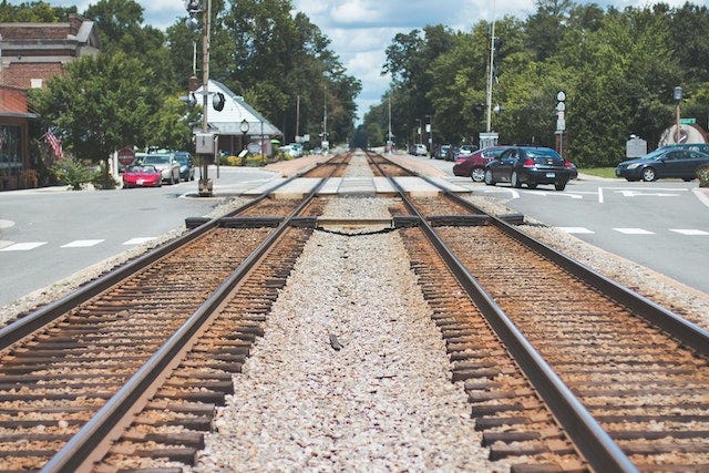 Railroad tracks run into the distance. A road in a town runs along both sides and cross the railroad at the center of the picture. There are parked cars on the left and driving cars headed toward the intersection on the right. Trees line the tracks going into the distance.