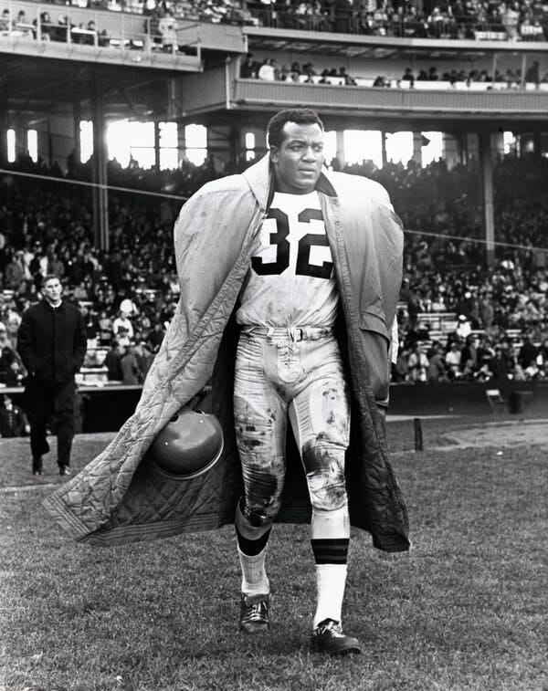 A black and white photo of Jim Brown, a muscular Black man with short hair wearing a No. 32 jersey and a coat, standing on the sidelines of a football stadium.