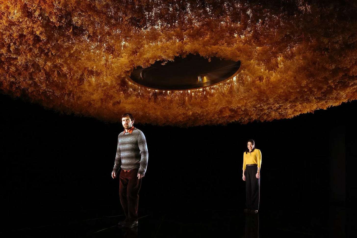 A photograph of a scene in the play "Constellations". Two actors stand on a dark black stage underneath a circular yellow floral ceiling piece. The character Roland wears a grey sweater and burgundy pants. The character Marianne stands behind him, grinning, wearing a yellow sweater and black pants.