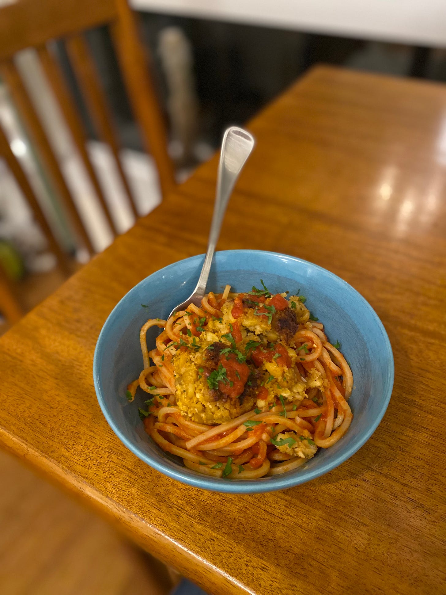 A blue bowl of spaghetti quadrato in marinara sauce, with two tempeh meatballs on top. One has broken apart into a few pieces. Everything is sprinkled with parsley.