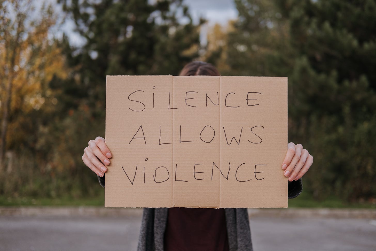 A person stands out side in the street holding up a cardbaord sign that says  Silence Allows Violence