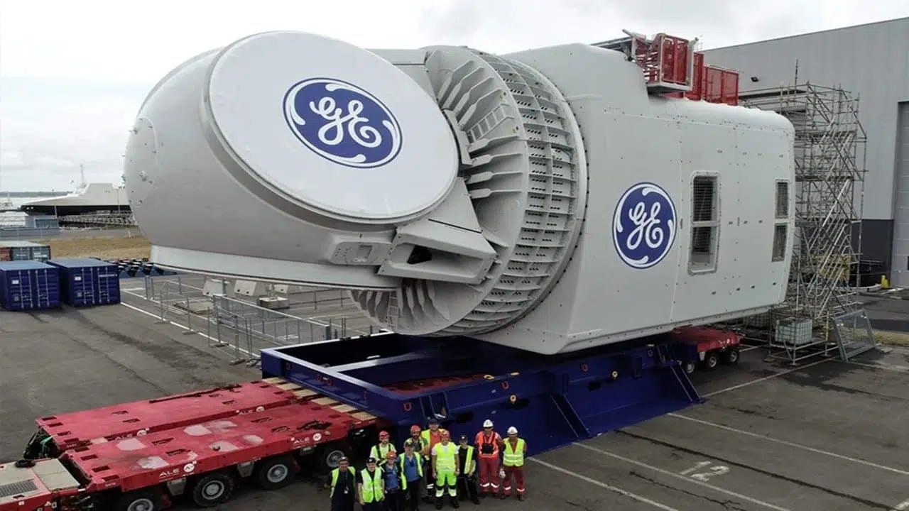 Foto de turbina gigante por trás de grupo de pessoas com capacetes e EPI. Elas ocupam 10% da foto. O restante é ocupado por esse imenso motor na cor branca com a logo azul da GE.