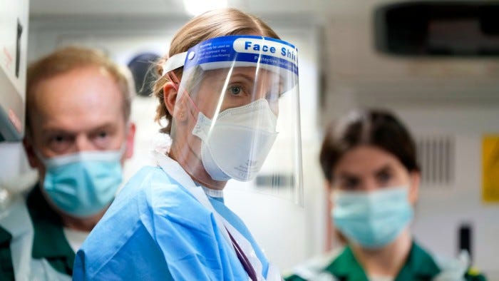 A female doctor in blue medical scrubs wearing a mask and face shield stands near two medical workers wearing face masks