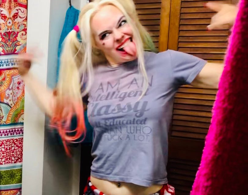 The author with tongue sticking out in a goofy grimace, pointing at herself while dressed as Harley Quinn in a t-shirt that says, "I am an intelligent, classy, educated woman who says 'fuck' a lot."