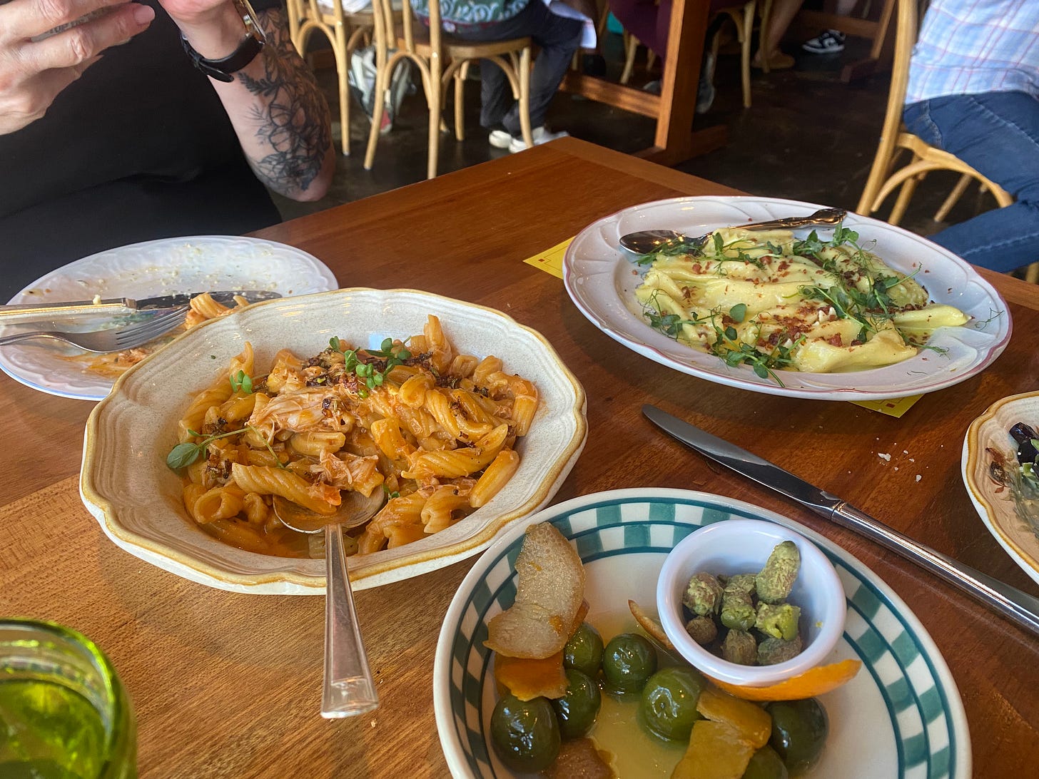 Several mismatched serving plates on a wood table, with side plates and utensils around. One has some castelvetrano olives with orange zest, some of the pits in a little dish. Another has the dungeness crab garganelli alla vodka, and the furthest from the camera has the agnolotti carbonara, covered with pea shoots.