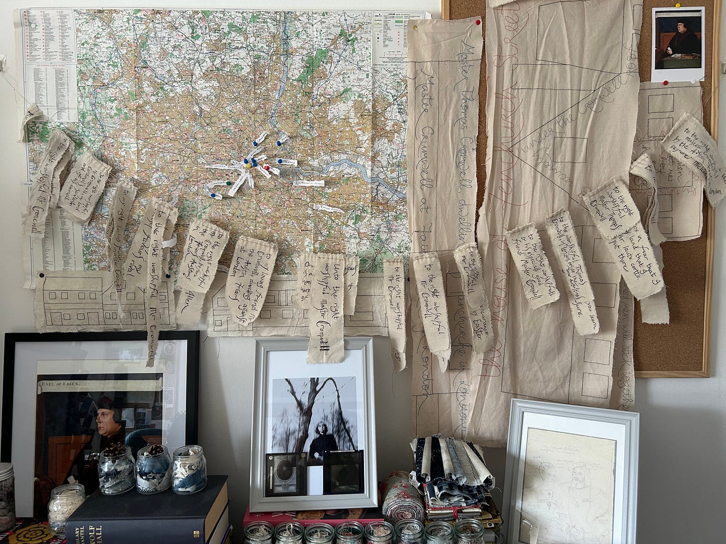 A map on a wall, a portrait of Thomas Cromwell, a photograph of Hilary Mantel, jars of thread and scraps of fabric. Hanging by the map is a row of fabric pieces, all of which feature inked writing representing the addresses of Thomas Cromwell.