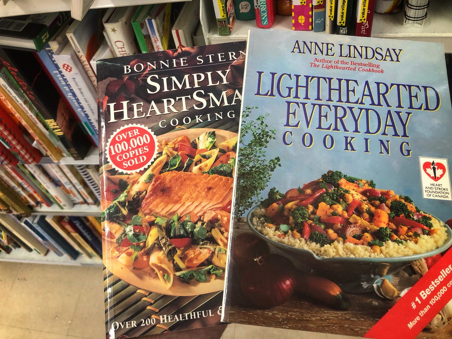 Copies of "Simply HeartSmart Cooking" by Bonnie Stern and "Lighthearted Everyday Cooking," by Anne Lindsay held up in front of some thrift store bookshelves
