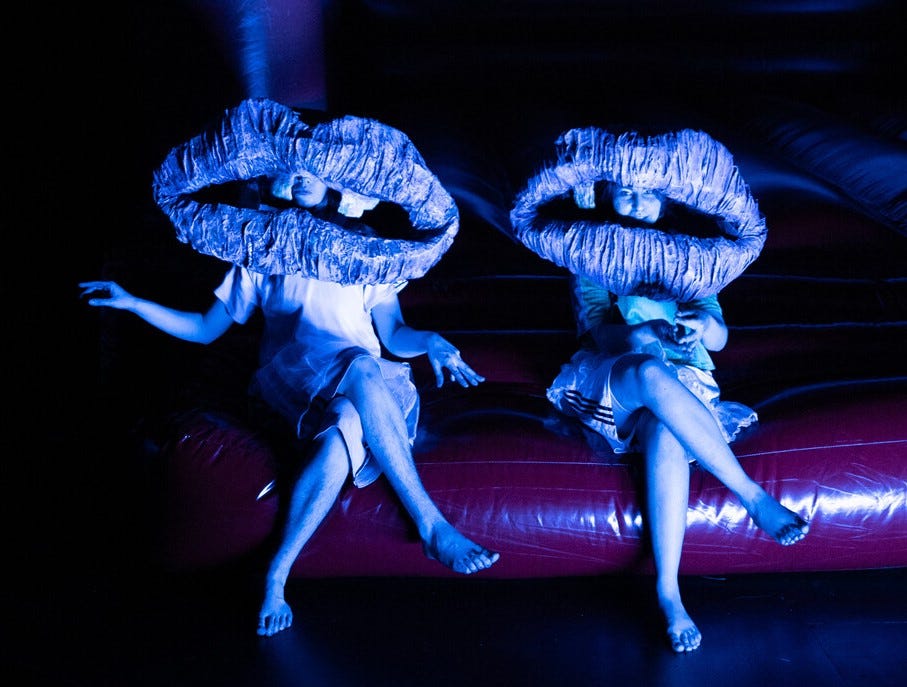 Die Schauspieler*innen Marie Schwanitz und Saba Hosseini tragen große Münder als Masken. Sie sitzen im Dunkeln, im Schein violetten Lichts am Fuß einer Hüpfburg. Eine Szene aus dem Theaterstück „Zertretung“ von Lydia Haider, inszeniert vom Schauspiel Stuttgart.