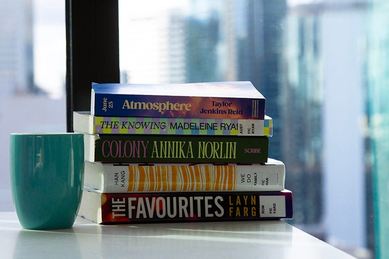 A stack of books next to a blue mug. The books are: ‘Atmosphere’ by Taylor Jenkins Reid, ‘The Knowing’ by Madeleine Ryan, ‘Colony’ by Annika Norlin, ‘We Do Not Part’ by Han Kang and ‘The Favourites’ by Layne Fargo. A stack of books next to a blue mug. The books are: ‘Atmosphere’ by Taylor Jenkins Reid, ‘The Knowing’ by Madeleine Ryan, ‘Colony’ by Annika Norlin, ‘We Do Not Part’ by Han Kang and ‘The Favourites’ by Layne Fargo.