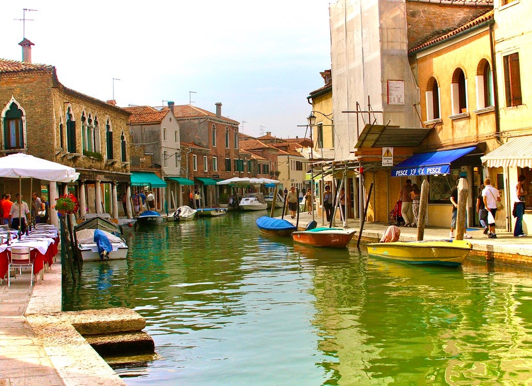 A canal with boats in the water with Murano in the background

Description automatically generated