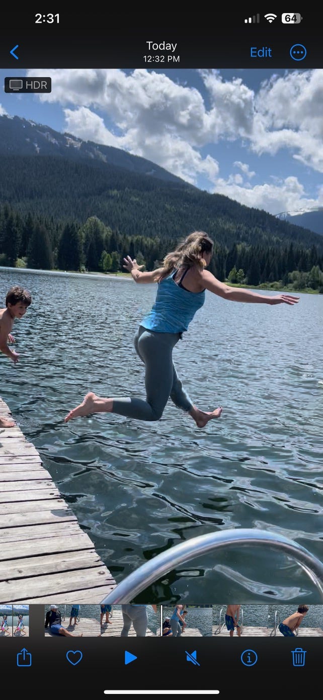 A clothed woman jumps off a dock into a lake while a child looks on.