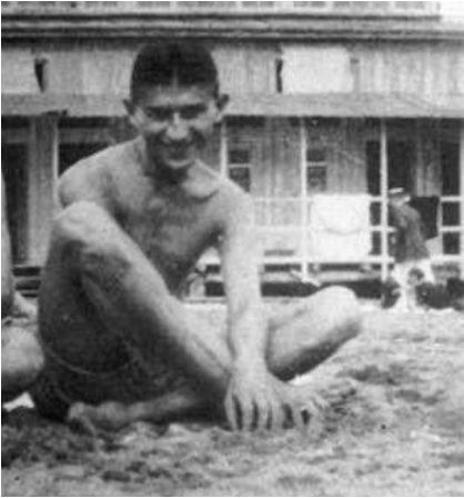 Thin young man sitting crosslegged at beach, grinning at camera