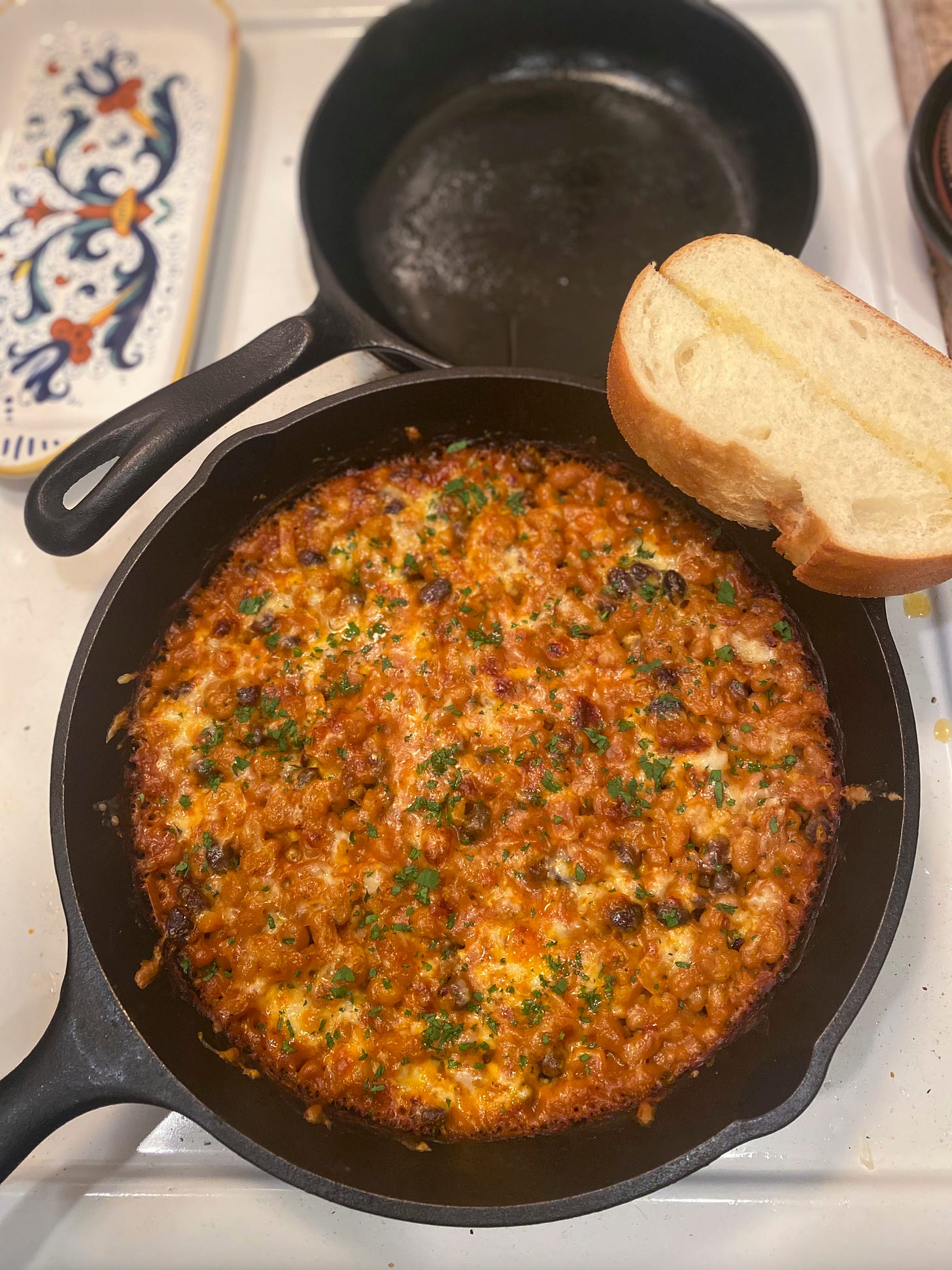 On top of the stove, a cast iron pan full of cheesy baked beans, sprinkled with parsley. A slice of garlic bread rests on the edge. Behind it is another smaller, empty cast iron pan, and a spoon rest with an artistic design in shades of blue, yellow, and orange.
