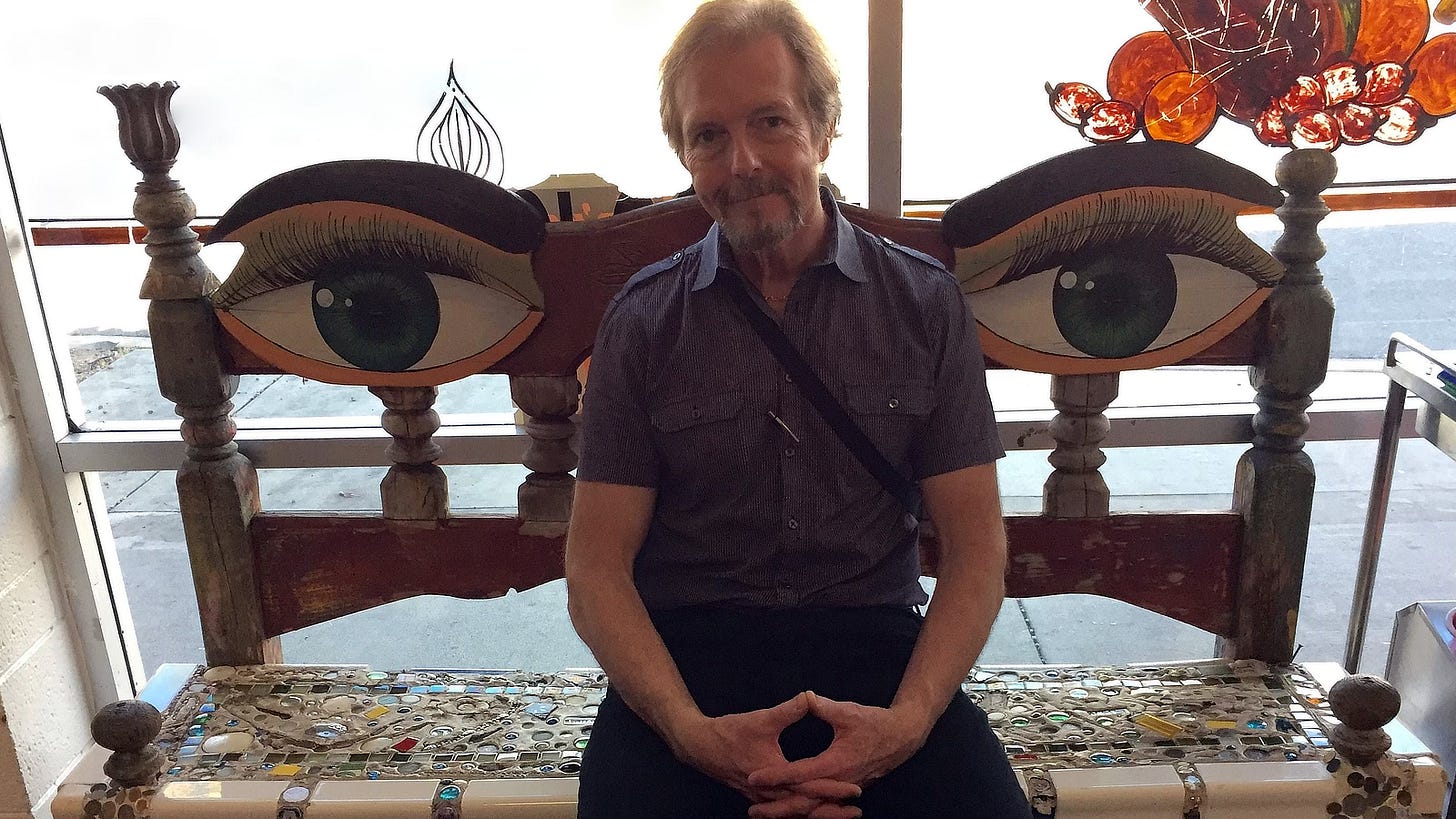 Artist Michael Whelan seated on a unique bench. The seat is a mosaic while the backrest is decorated with two large human eyes flanking either side of Michael. His fingers are interlaced with thumbs touching and his cheeks flare with dimples.