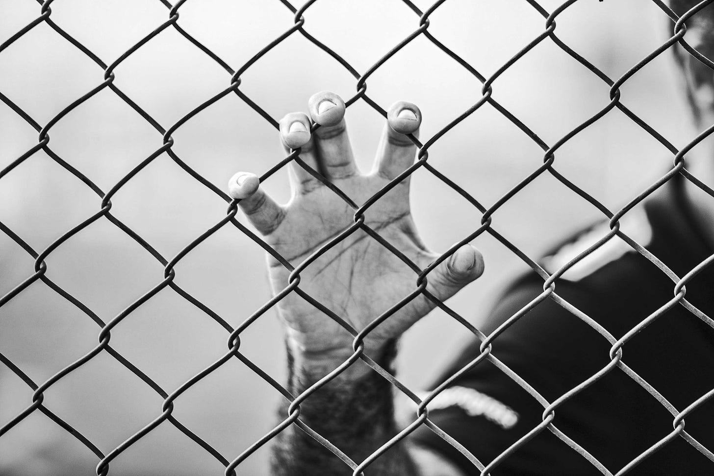 A man stands behind a chain link fence. He is blurred in the background while the focus is on his hand grasping the chain link fence. 