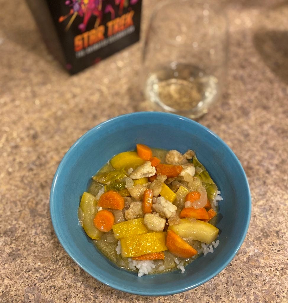 A blue bowl of the green curry described above, pieces of yellow zucchini, carrot, and tofu crumbles throughout with a few basil leaves, all over jasmine rice. In the background is a glass of white wine (and the popcorn box from the Star Trek event but never mind that).