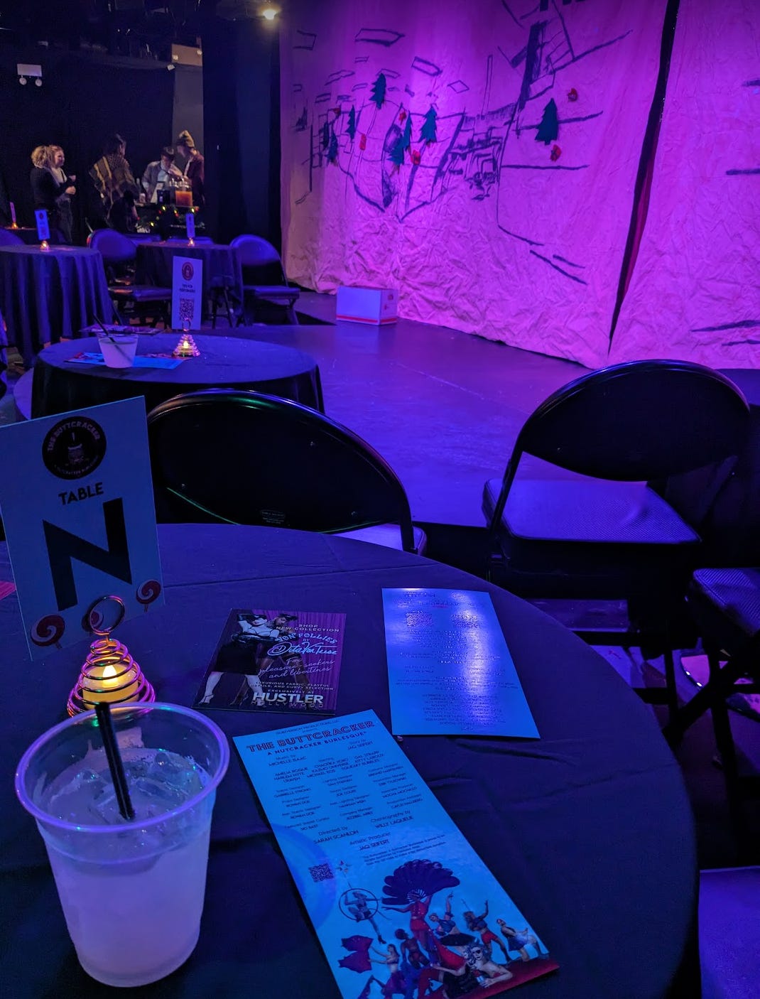 A purple light dimmed stage sits across from foldable black chairs. There is a margarita drink in a plastic cup on a table next to the flyer for the Buttcracker show