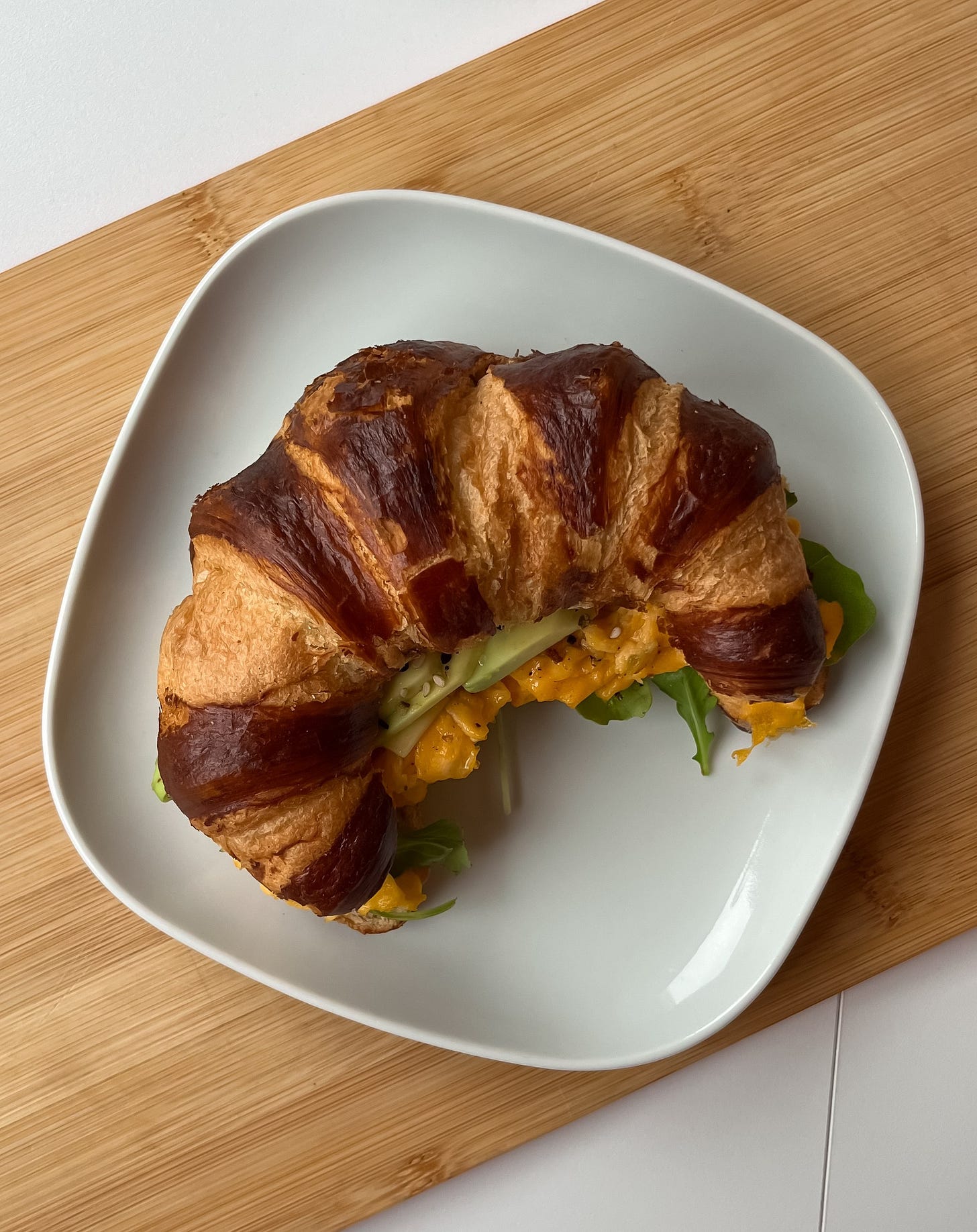 Close-up of a warm croissant stuffed with fried eggs, avocado slices, melted cheese, and fresh arugula, sprinkled with everything bagel seasoning