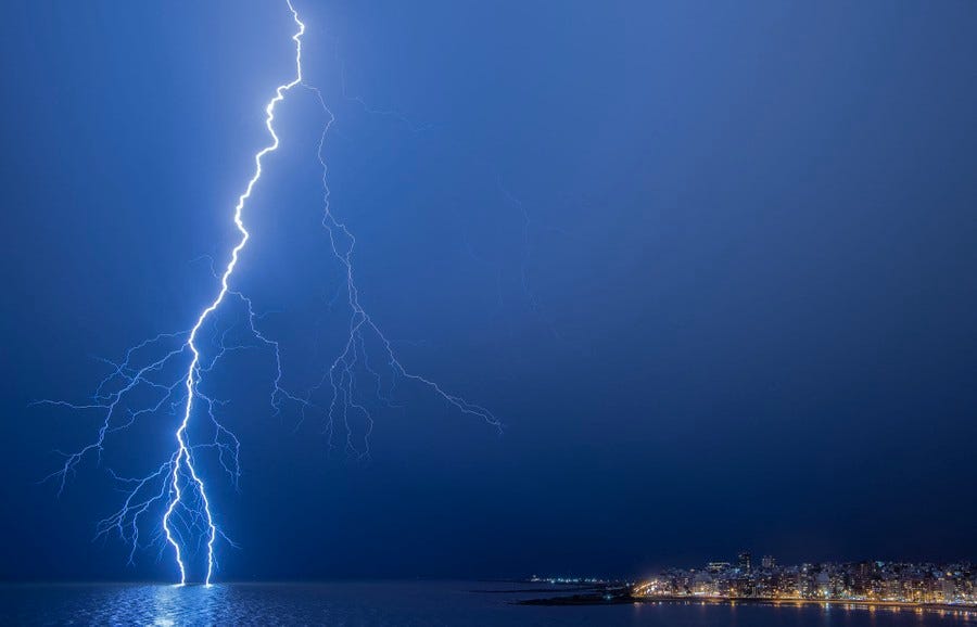 Lightning strikes the ocean's surface near a city, captured from a distance.