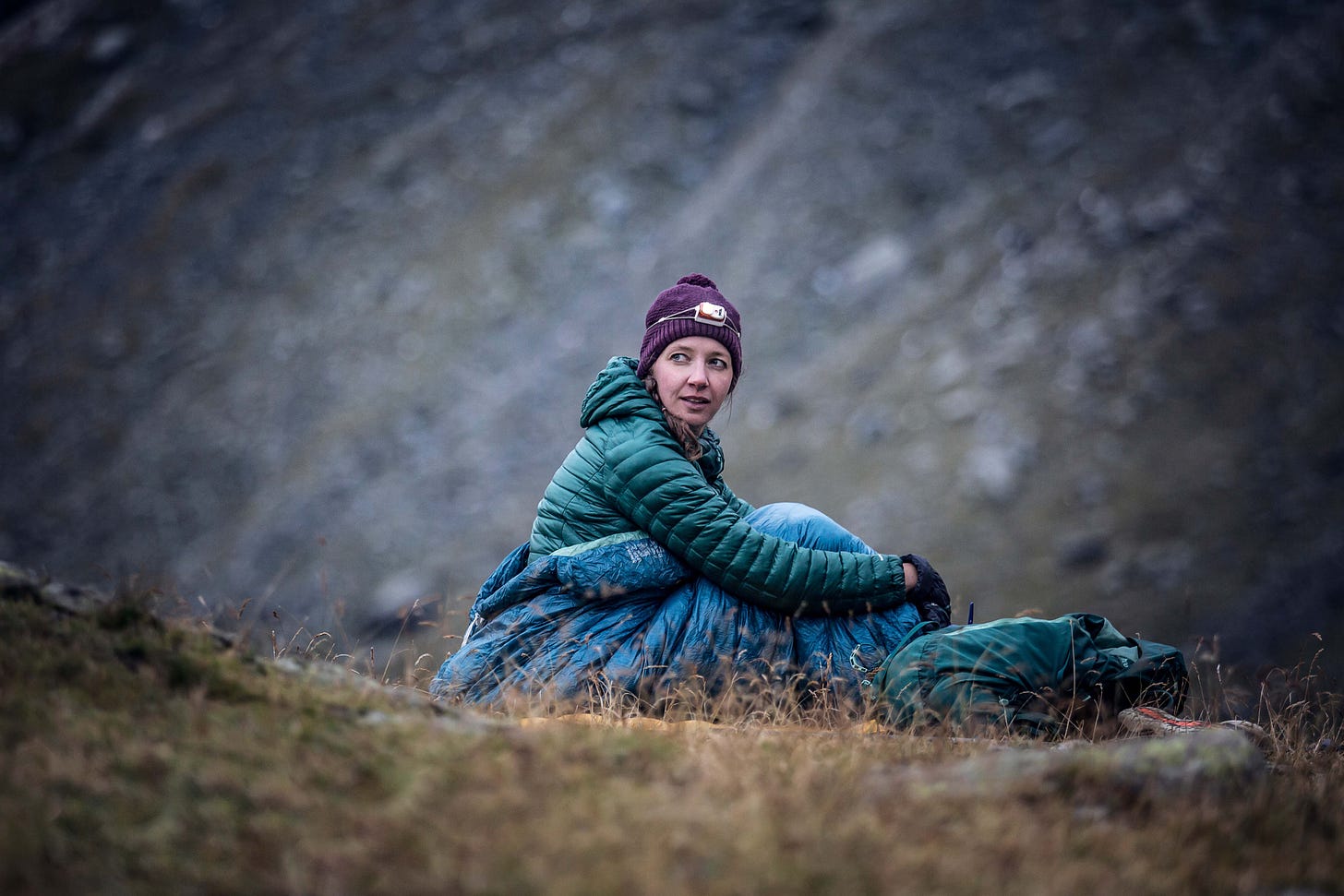 Jenny sits alone in her sleeping bag on dry brown grass, she wears a bobble hat with headtorch, down jacket and gloves