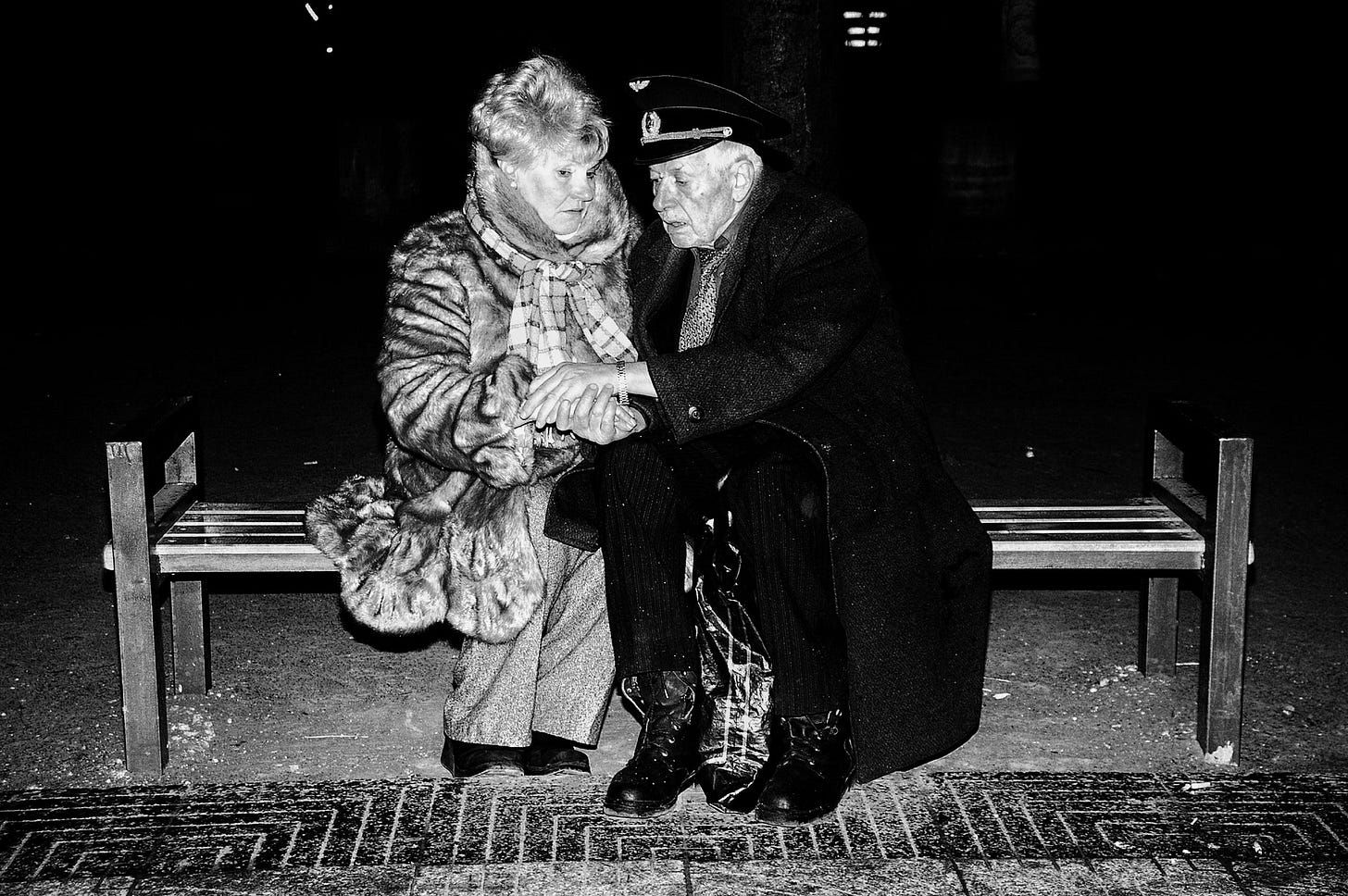 Elderly couple holding hands on a bench at night.