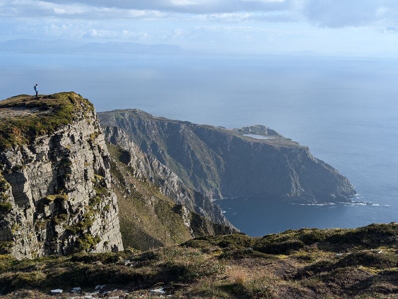 A person stands over a sun-kissed precipice looking out to the sea. There are cliffs in the shadows behind, dropping into the sea. A cliff-top lake just catches the sun above these dark cliffs. There is land visible in the far distance, on the other side of a wide expanse of sea. There are a few, high fluffy clouds in the background.