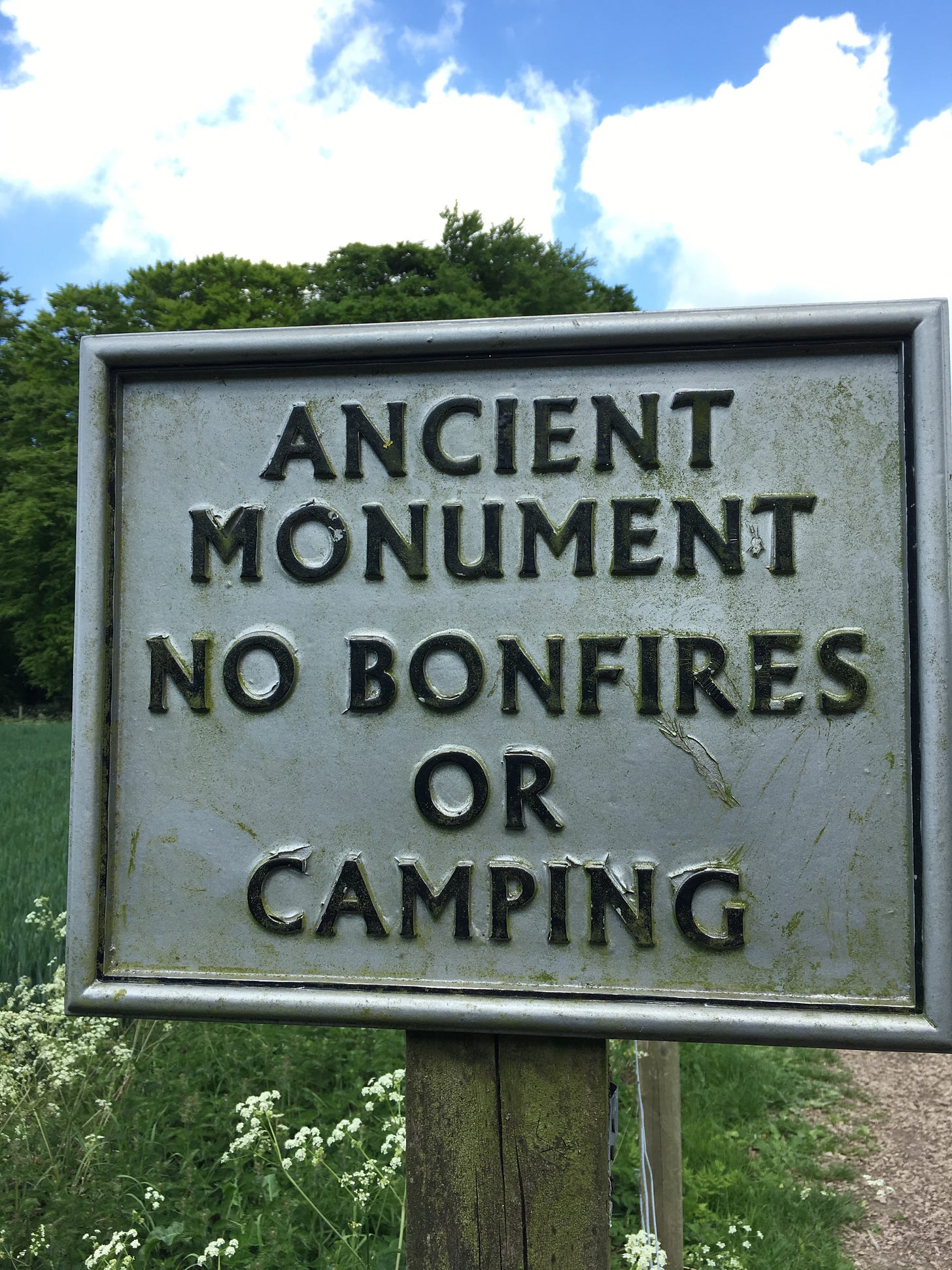 a sign the reads "ANCIENT MONUMENT NO BONFIRES OR CAMPING," with flowers and trees nearby.