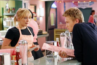 Still from A Cinderella Story where Sam is behind the counter in her pink diner apron, waiting on Austin who's holding a menu and about to order.