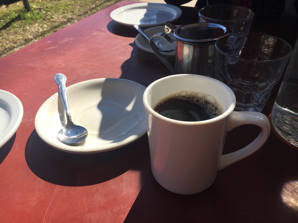 A red table in the sun is covered in plates, utensils, cups, and a single cup of black coffee