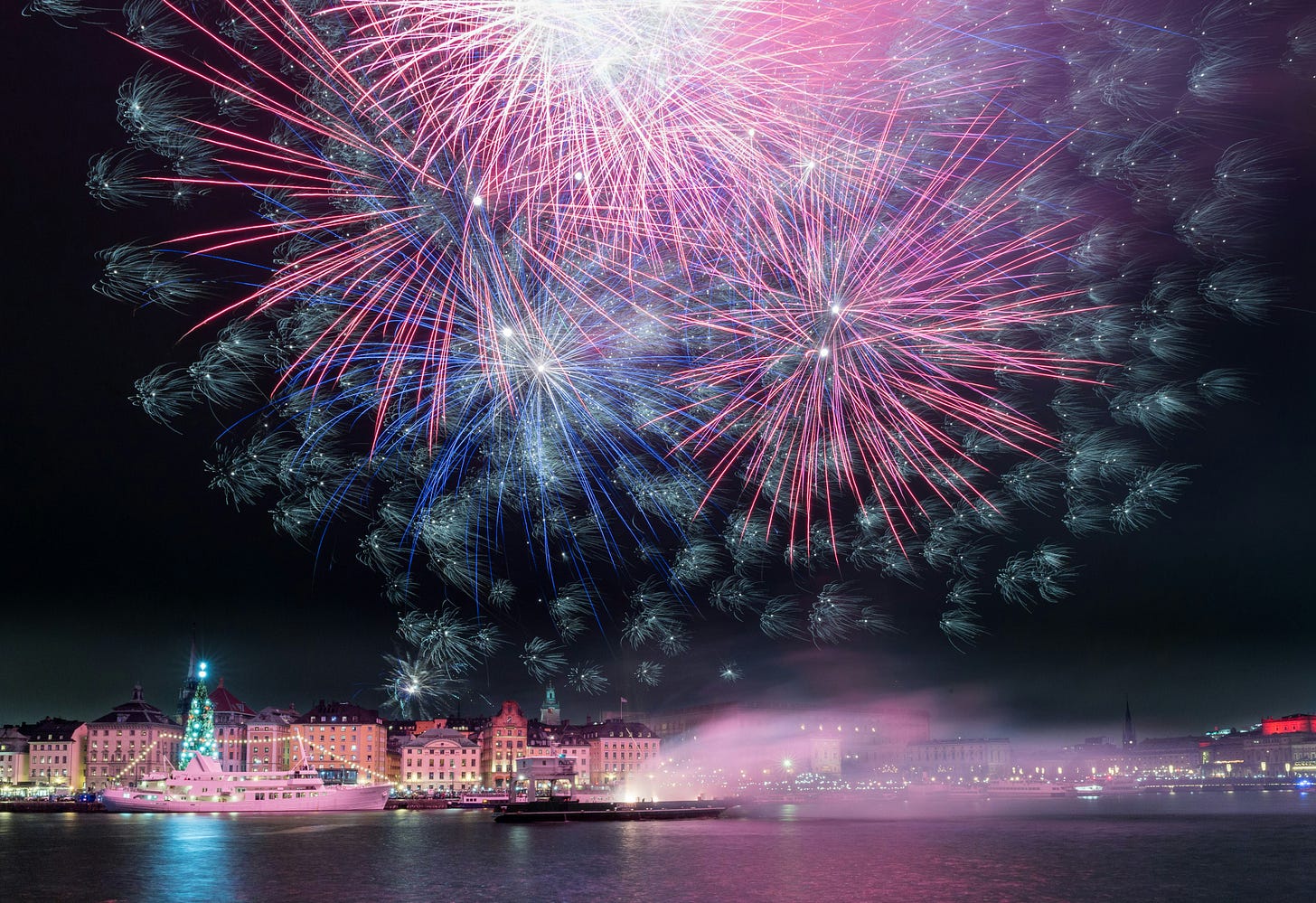 A big fireworks display over a historical Old Town with a brightly lit Christmas tree