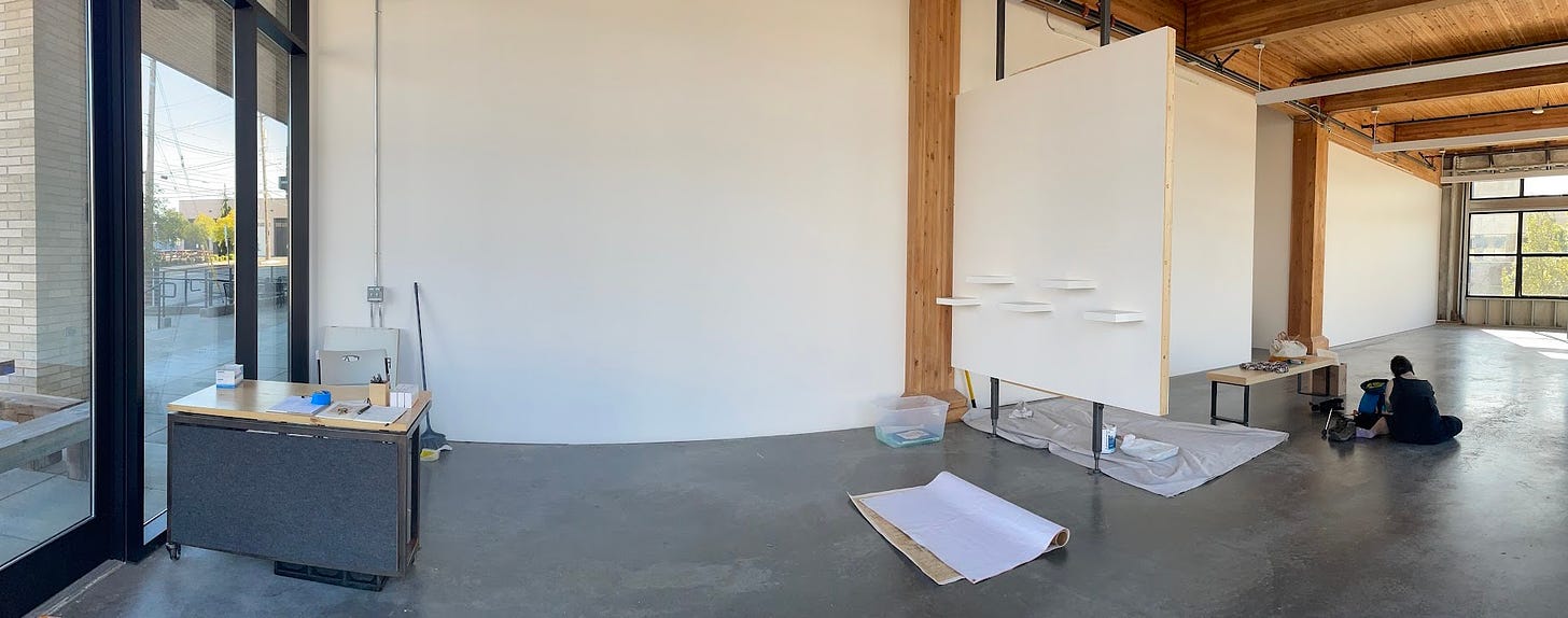 panoramic image of a gallery space; a moveable desk is positioned at lower left, with views of a long white wall, a smaller movable white wall, and an artist sitting on the floor working
