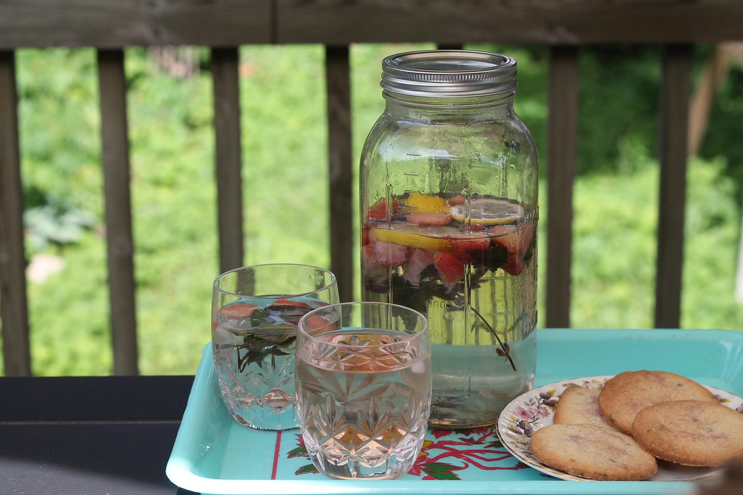 A large mason jar containing water with fruits and herbs sits beside two glasses and a plate of shortbread cookies