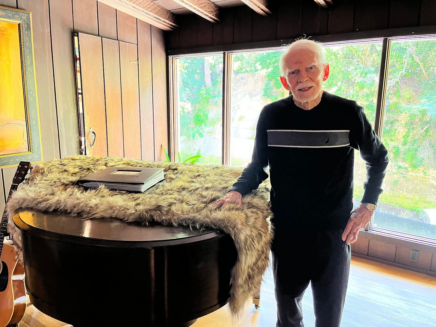 Elderly man with black and white track suit stands in front of piano. The room is paneled wood. Floor to ceiling windows show greenery. A guitar rests on the floor near the piano. The piano has a blanket lying over its top.