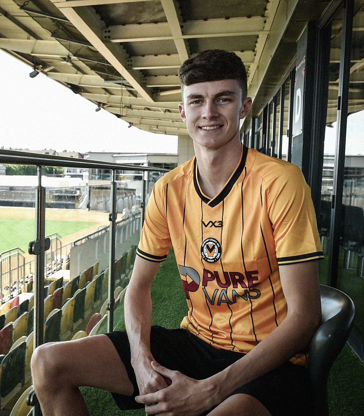 A photo of Joe Thomas in an orange Newport County shirt sat on a balcony at Rodney Parade