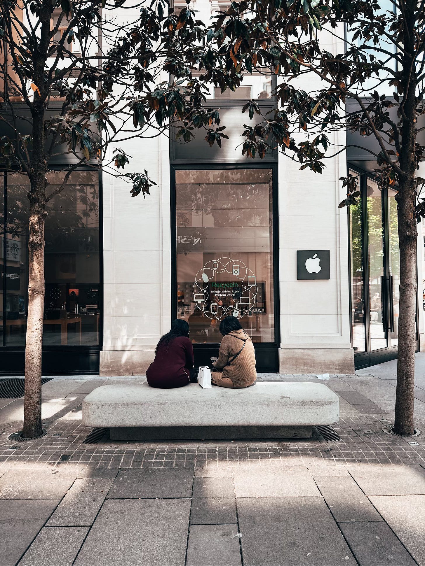 Two people sit on a stone bench outside the Apple Store in Vienna. On the window is an Earth Day recycling decal.